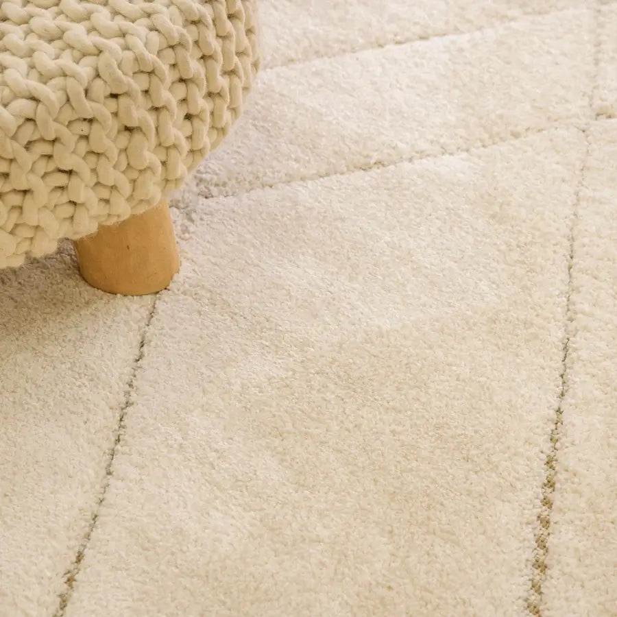 Close-up of a cream-colored shag rug with stitched geometric lines, showing a chunky knit stool leg on the left.