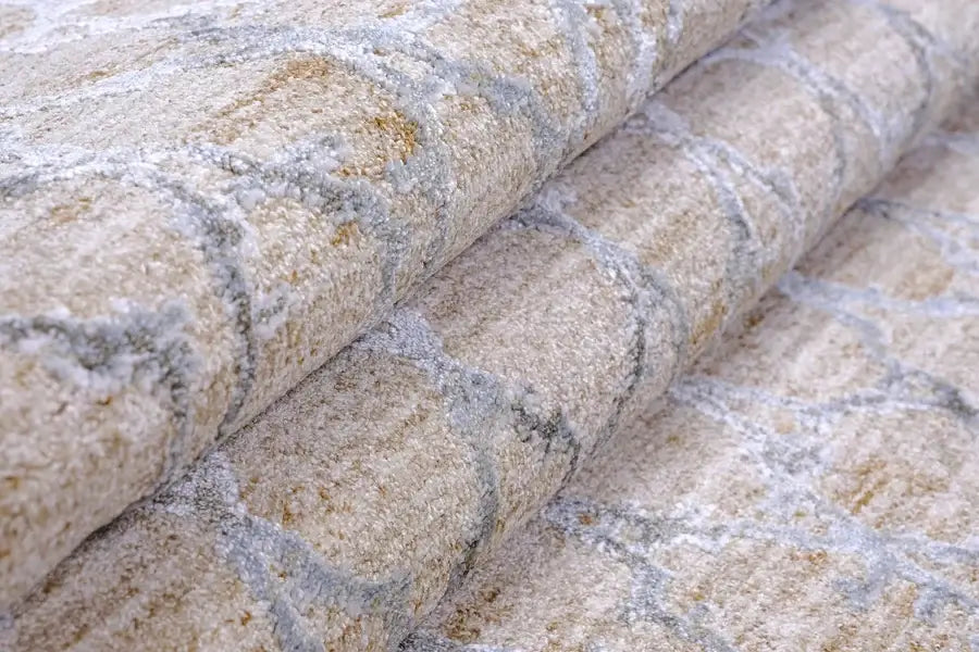 A close-up of a folded area rug with a textured, shaggy surface in beige and cream tones, featuring a gray geometric trellis pattern.