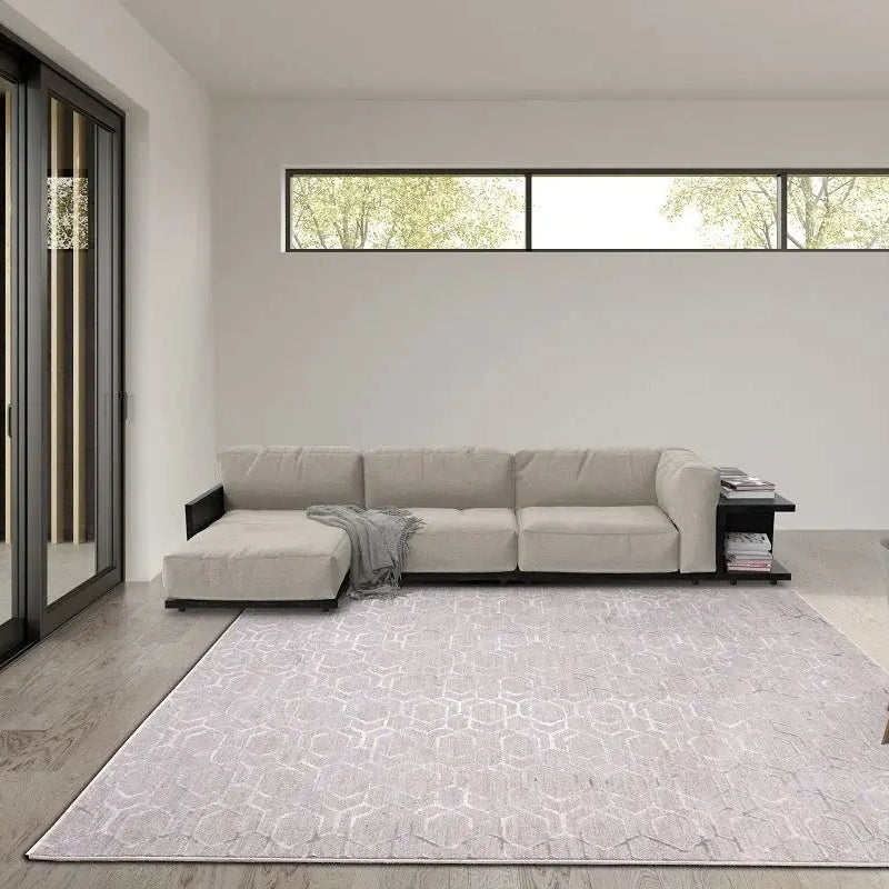 A large rectangular area rug with a light gray and white geometric pattern is displayed in a modern living room setting, positioned in front of a beige sectional sofa.
