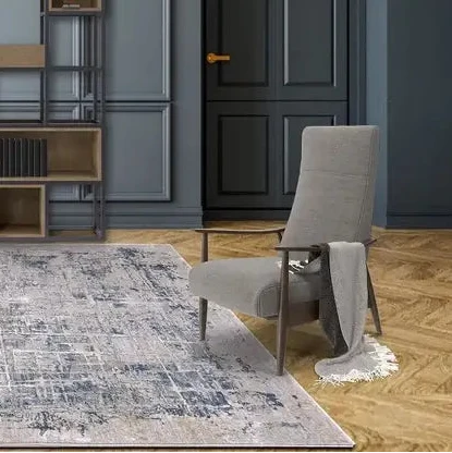 A modern living room scene featuring a distressed gray and blue area rug on a light wood floor, with a gray upholstered armchair, a tall black metal bookshelf, and a dark paneled door in the background.