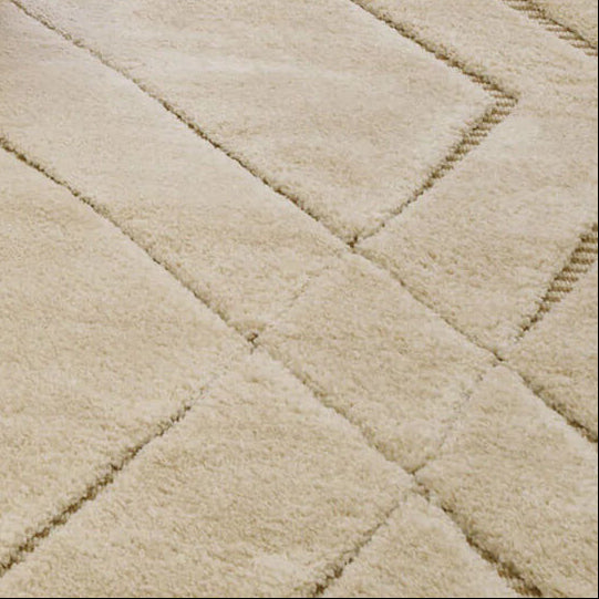 A large cream-colored area rug with a raised geometric diamond pattern, styled in a cozy living room setting with a chunky knit ottoman and a leather chair.