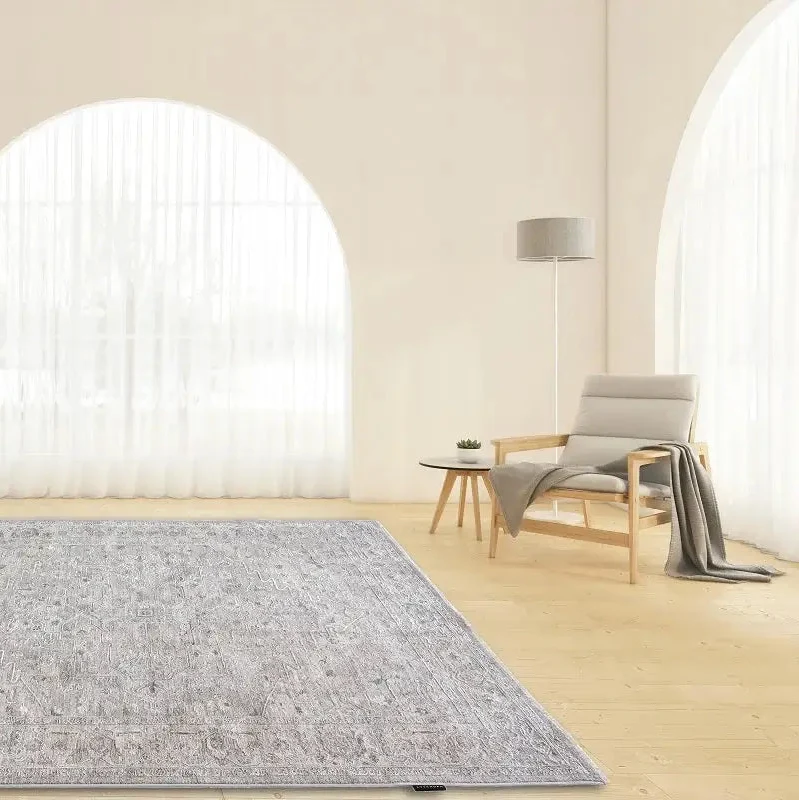 A rectangular area rug with a distressed, faded traditional pattern in shades of gray and white, displayed on a light wood floor in a bright, modern room with arched windows and a minimalist armchair.