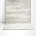 A textured area rug with an abstract wavy pattern in shades of gray, beige, and off-white, draped against a white brick wall and white floor.