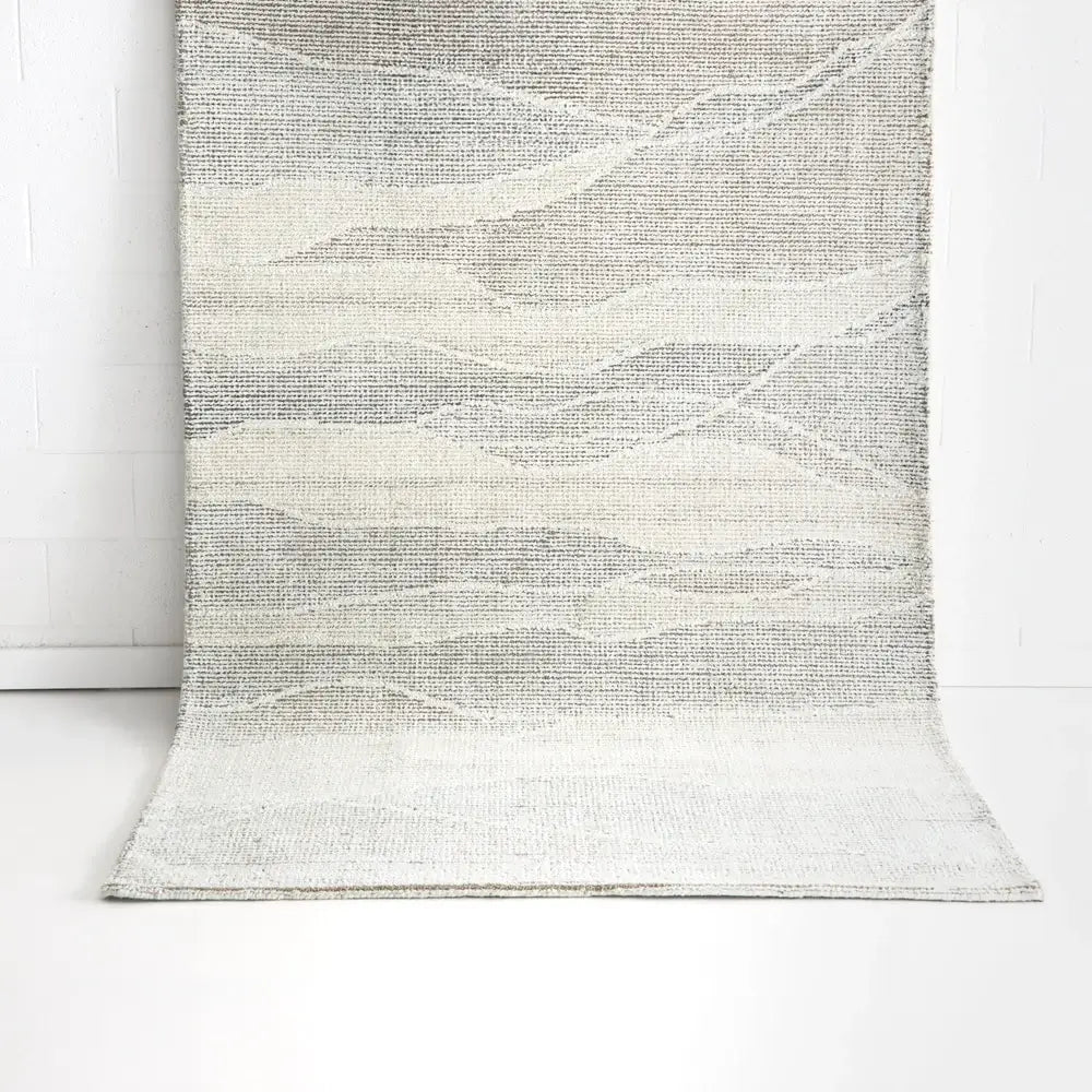 A textured area rug with an abstract wavy pattern in shades of gray, beige, and off-white, draped against a white brick wall and white floor.
