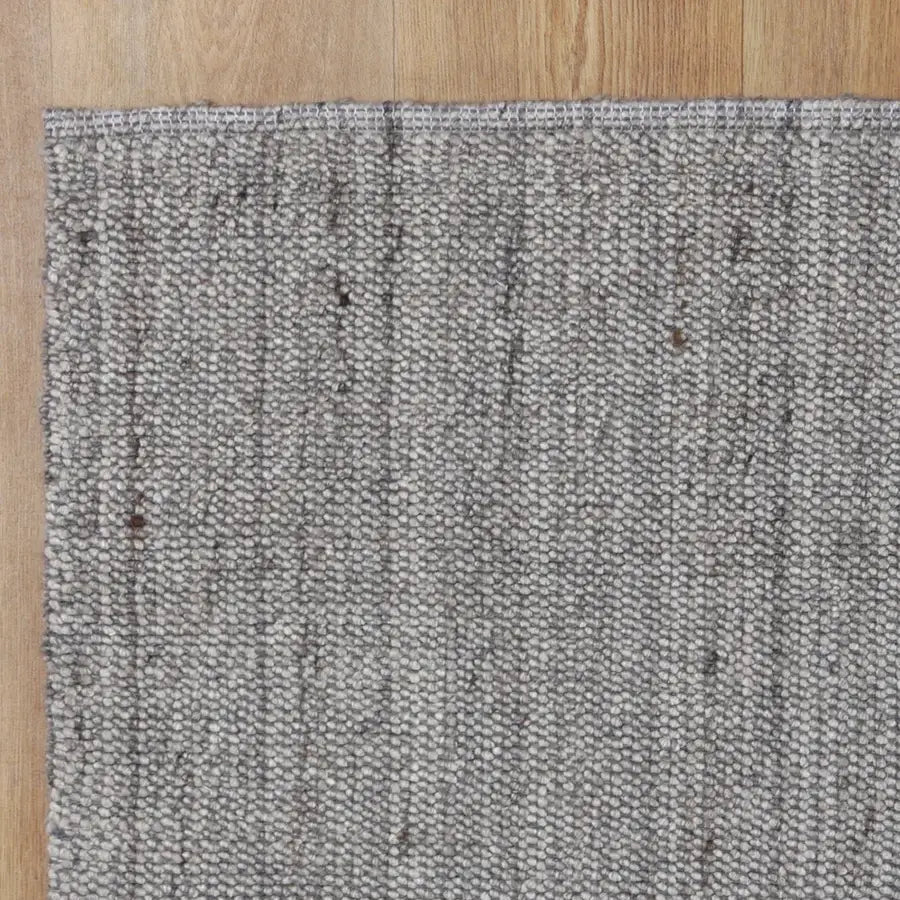 A close-up of a textured woven area rug in a heathered gray and white tone, shown on a light wood floor.