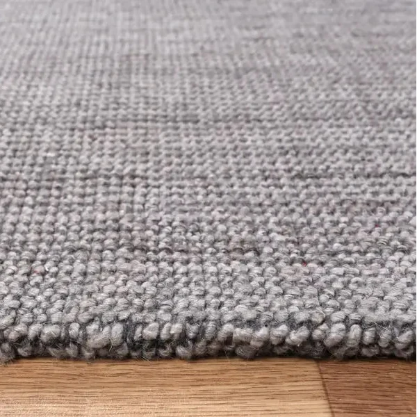 Close-up of a gray woven rug with a textured, chunky knit pattern and a frayed edge, shown resting on a light brown wooden floor.